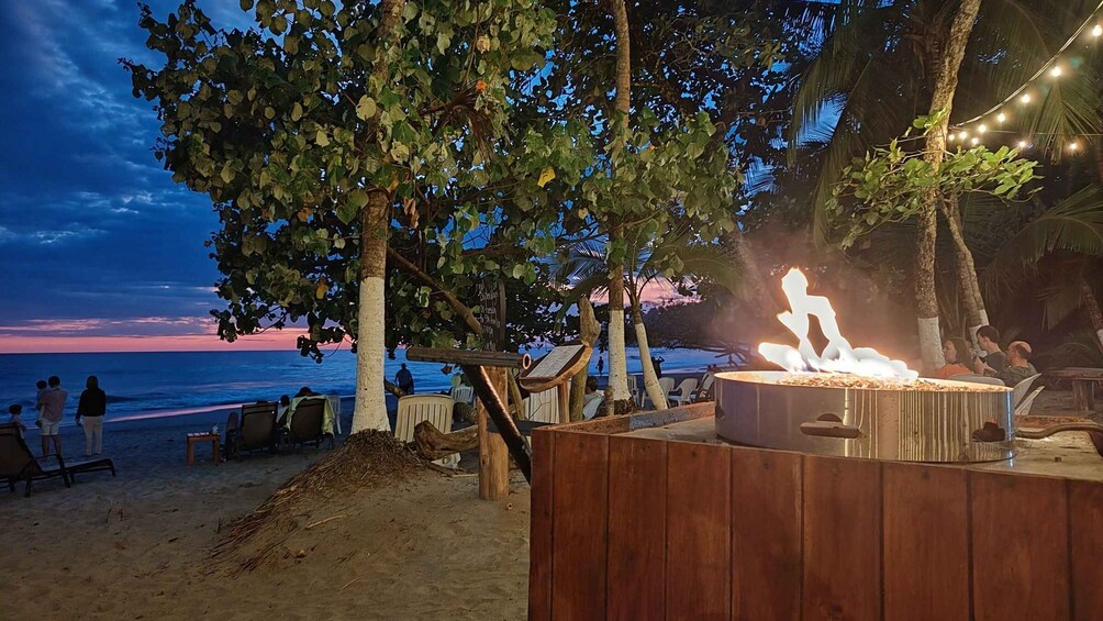 A beachside barbecue area with a fire pit, wooden seating, and a view of the ocean at sunset. 3