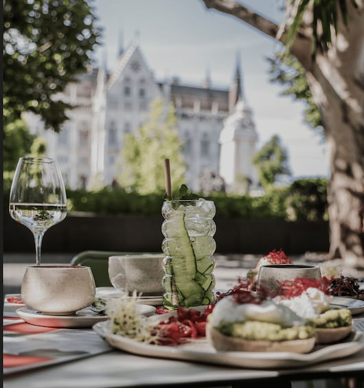 A table with a glass of white wine, a cucumber drink, and various food items. 1