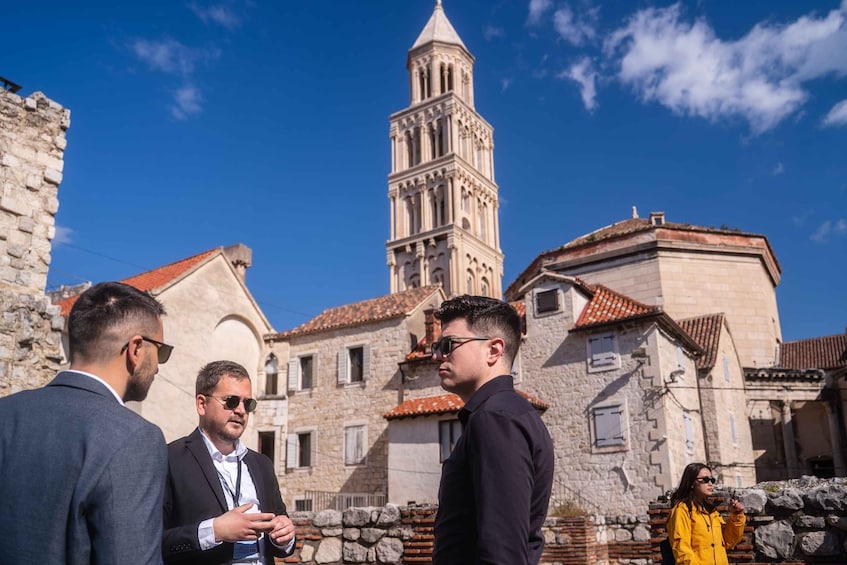 A group of people standing in a historic town square with a prominent tower in the background. 1