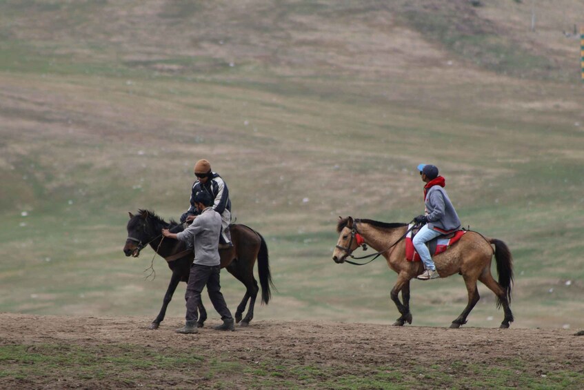 Two people leading horses on a dirt path in a hilly area. 1