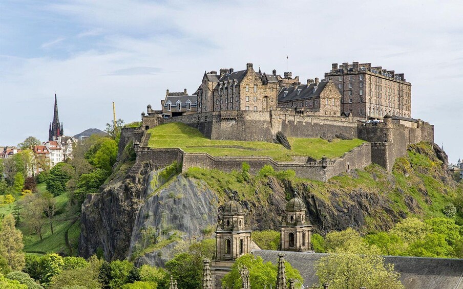Edinburgh Castle atop a hill with surrounding greenery and a cathedral in the background. 1