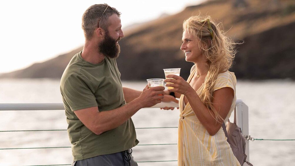 A couple holding drinks by a railing with a body of water and hills in the background. 1