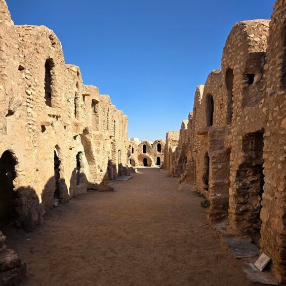 An ancient stone pathway flanked by ruins with arched doorways, leading towards a distant structure. 2