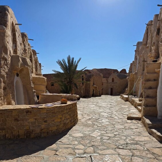 A stone-paved courtyard with traditional architecture, palm trees, and a clear blue sky. 5