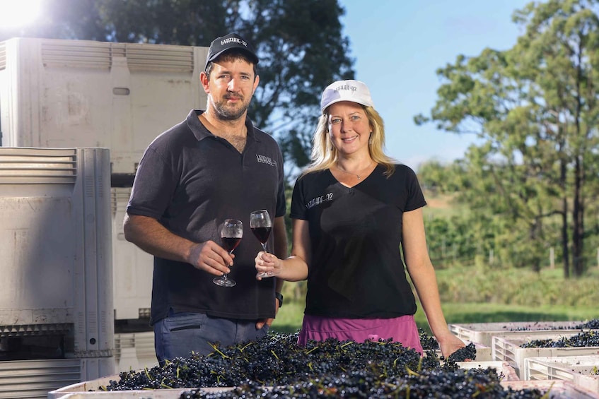 Two people holding wine glasses in a vineyard with grape clusters and a large industrial machine in the background. 4