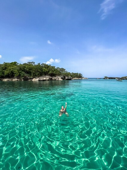 A person swimming in clear turquoise water with a rocky coastline and trees in the background. 2