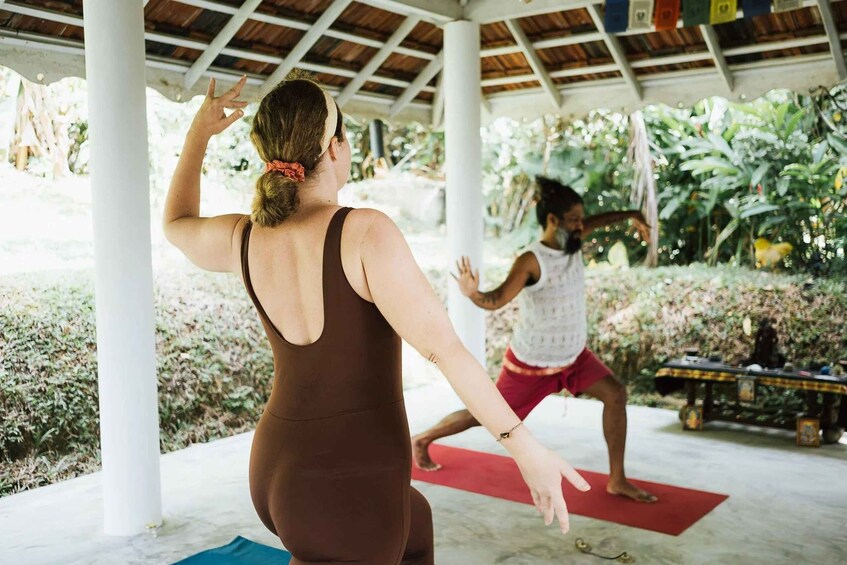 Two people practicing yoga outdoors under a covered area with greenery in the background. 2