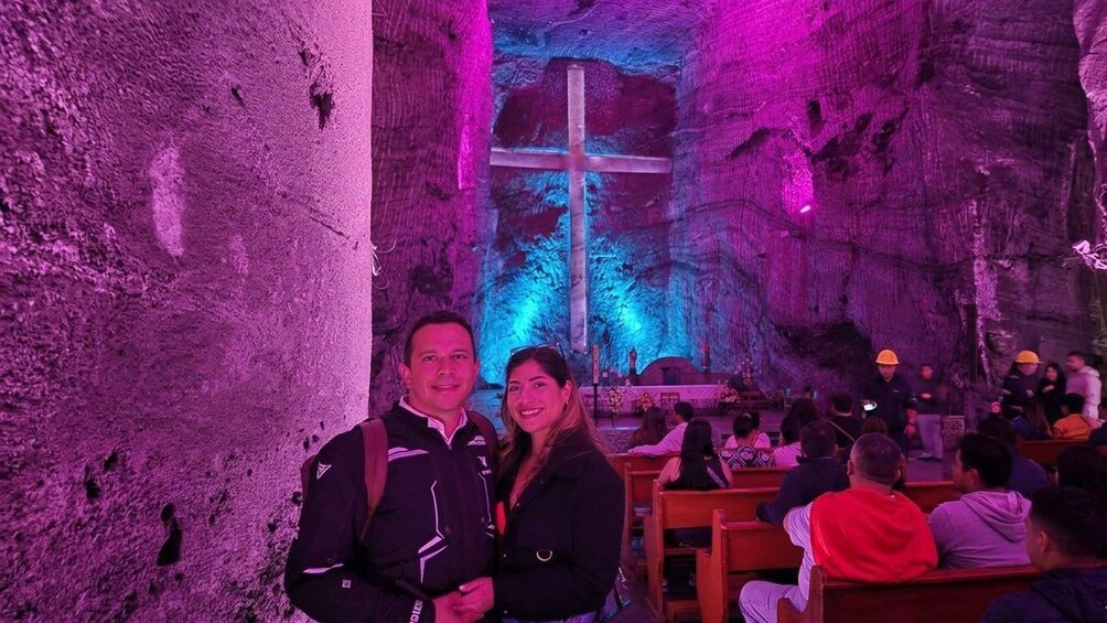 A cave interior with a cross illuminated in the center, people seated on benches, and a man and woman standing in the foreground. 4