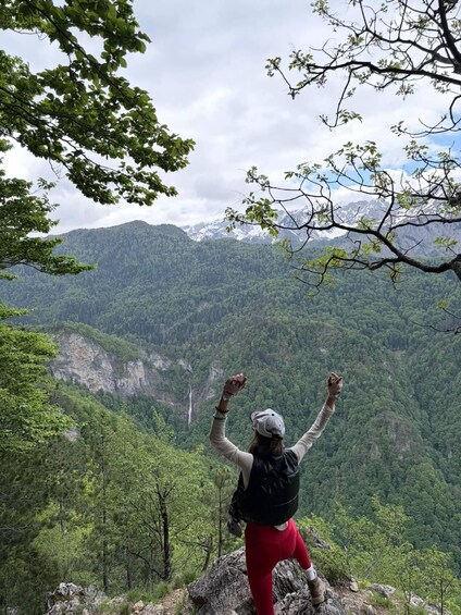 A person standing on a rocky outcrop with arms raised, overlooking a mountainous landscape with dense forests and a waterfall. 3