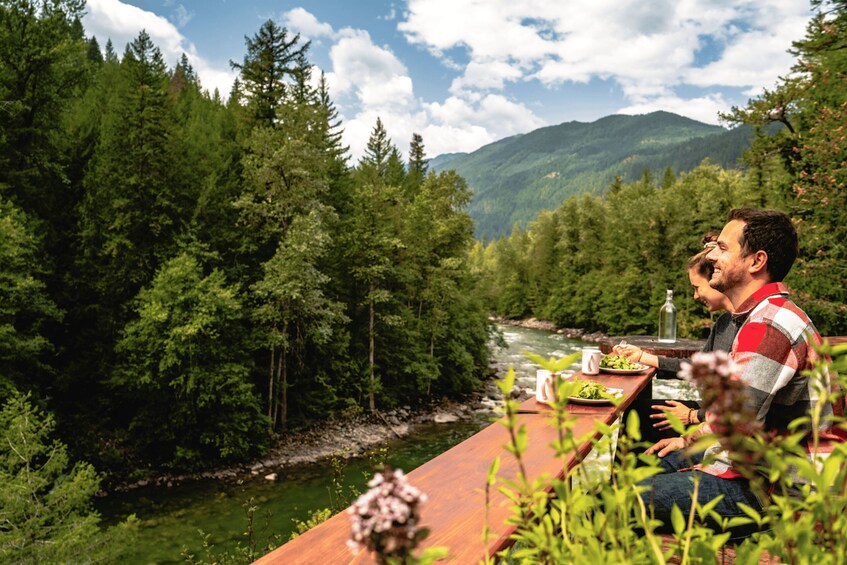 A man sitting at a wooden table by a river, surrounded by dense forest and mountains in the background. 2