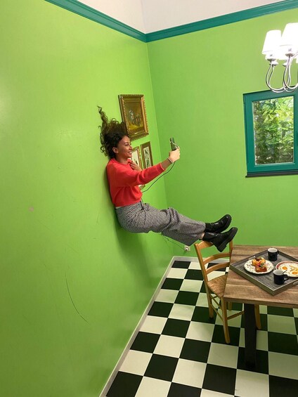 A person leaning against a green wall, holding a device, with a checkered floor and a table with food and drinks. 5