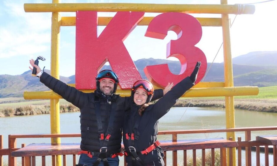 Two people with arms raised in front of a large red 'K3' sign, wearing helmets and harnesses, with a lake and mountains in the background. 4