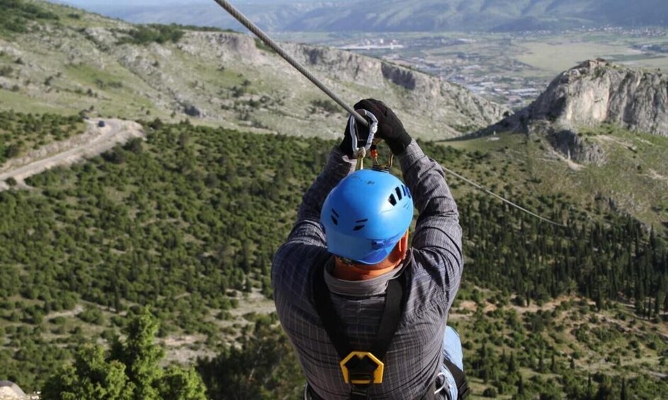 A person in a blue helmet and gray jacket, holding onto a zip line, overlooking a mountainous landscape with dense forests and rocky terrain. 1