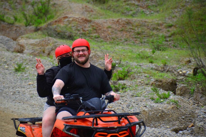 Two people on an orange ATV with a side cargo box, wearing red helmets, in a rocky outdoor setting. 2
