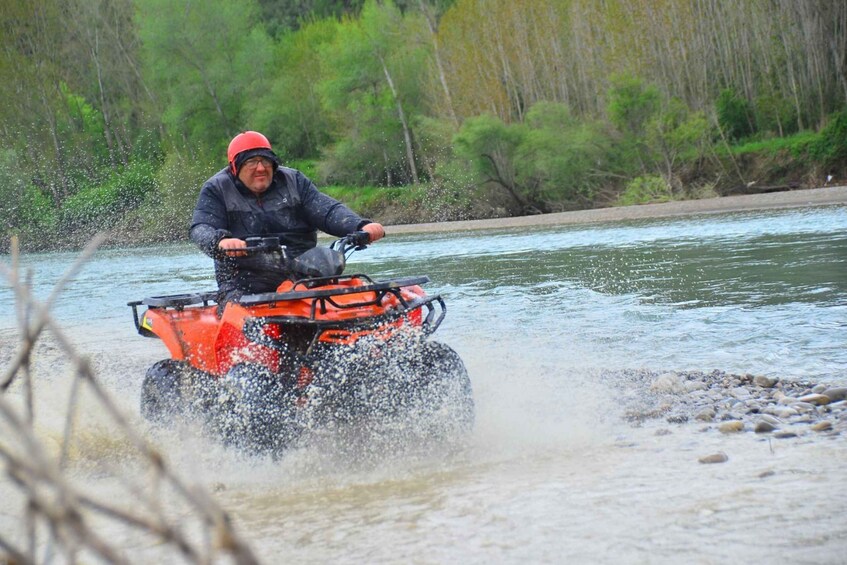 A person riding an ATV through a river, surrounded by trees and rocks. 1