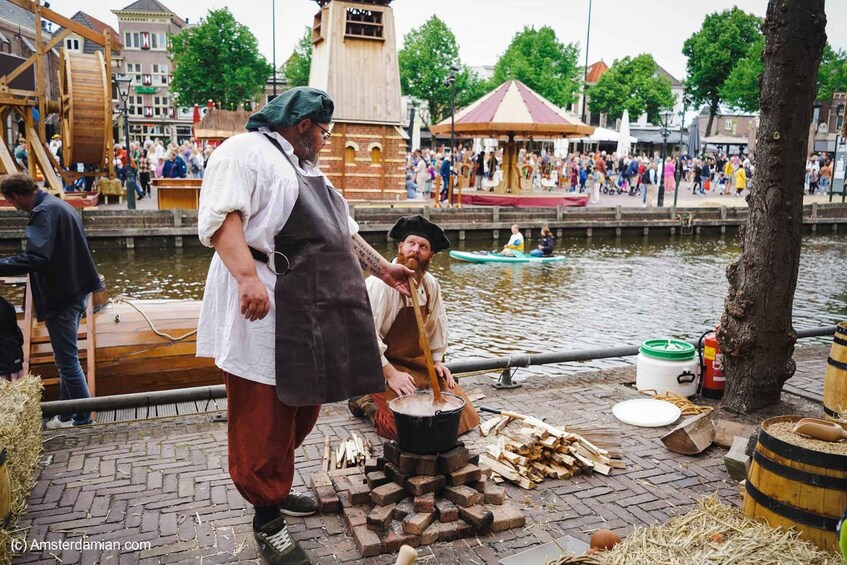 A person cooking outdoors by a river, with a wooden structure and a crowd in the background. 4