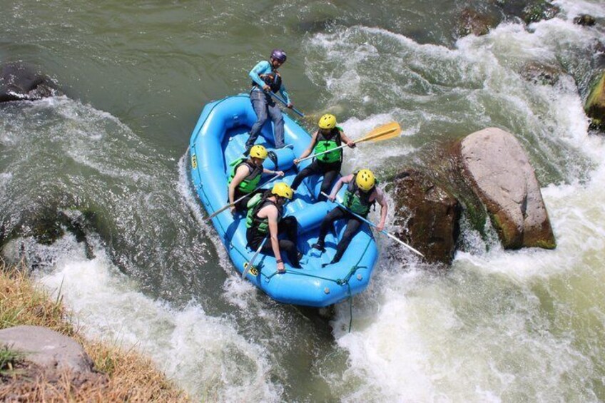A group of people rafting on a blue inflatable raft navigating a river with rapids and rocks. 3