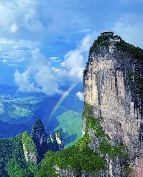 A towering cliff with a structure on top, a lush green forest, and a visible rainbow. 3