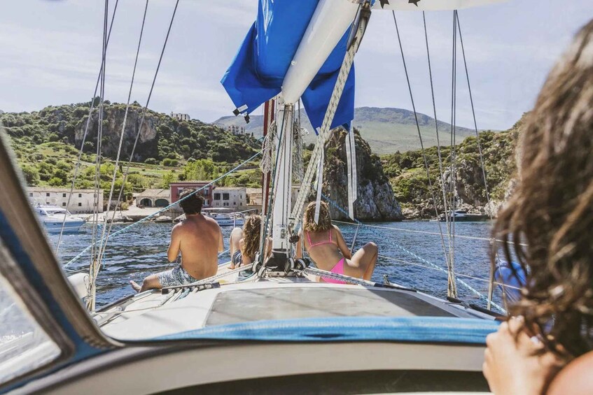A sailboat with people on board, a rocky coastline, and a building in the background. 5