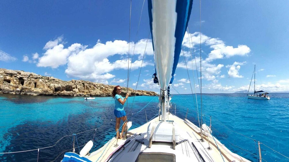 A sailboat with a person on deck, clear blue waters, and rocky cliffs in the background. 1