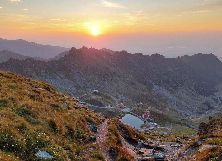 A mountainous landscape with a winding road, a small settlement, and a lake at sunset. 1