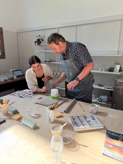 Two people in a pottery studio with clay, tools, and a magazine on the table. 1