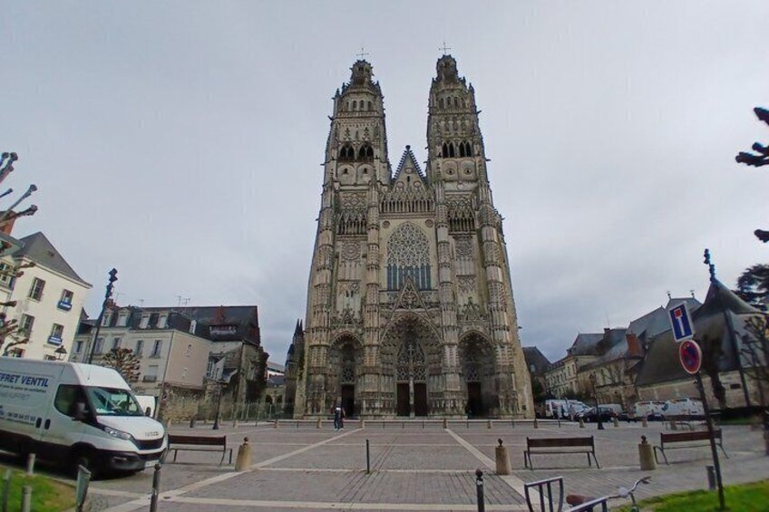 A Gothic cathedral with twin towers, a paved square in front, and parked cars. 1