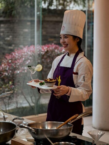 A chef holding a plate with a dessert and a glass of drink, with cooking utensils and a window with pink flowers in the background. 1