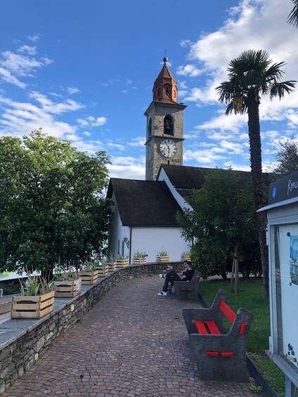 A cobblestone pathway leading to a church with a clock tower, surrounded by greenery and benches. 1