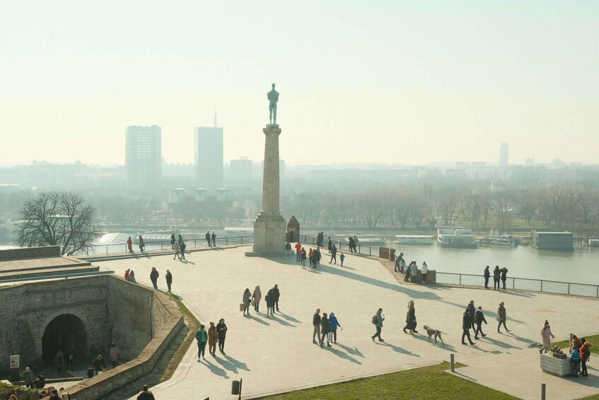 A public square with a monument, people walking, and a river in the background. 3