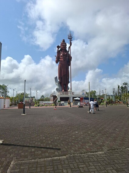A large statue of a deity holding a trident, with people walking around and a building in the background. 5