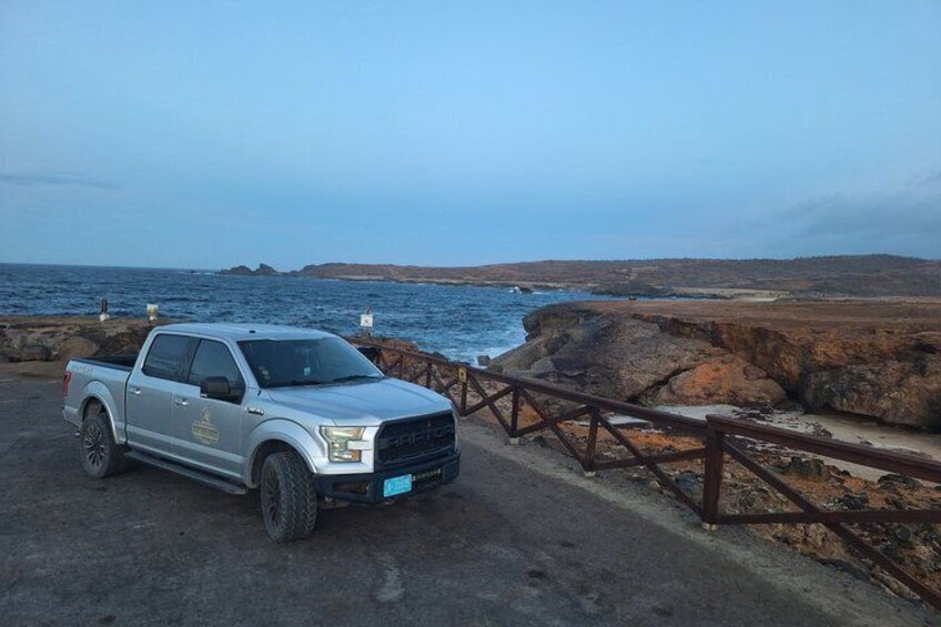 A silver pickup truck parked on a coastal road with a wooden fence and rocky terrain in the background. 3