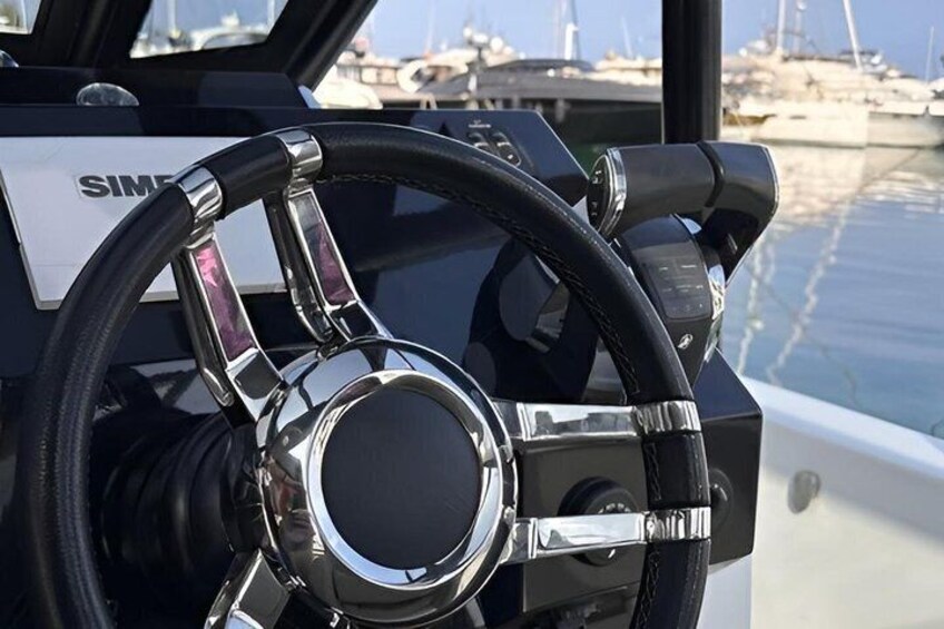 A boat's steering wheel with a SIMS logo, a chrome steering wheel, and a marina in the background. 2