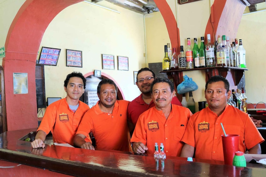 A group of people in orange shirts behind a bar counter with various bottles and a red cup. 5