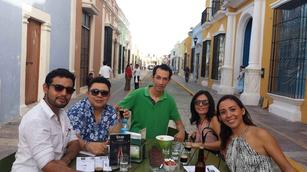 A group of people sitting at a table on a street with colorful buildings. 1