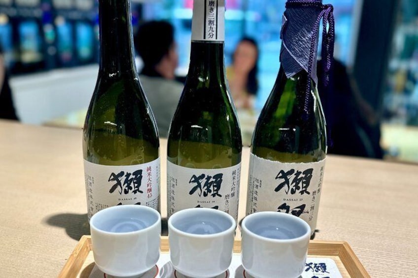 Three bottles of sake with white labels and three small white cups on a tray. 3