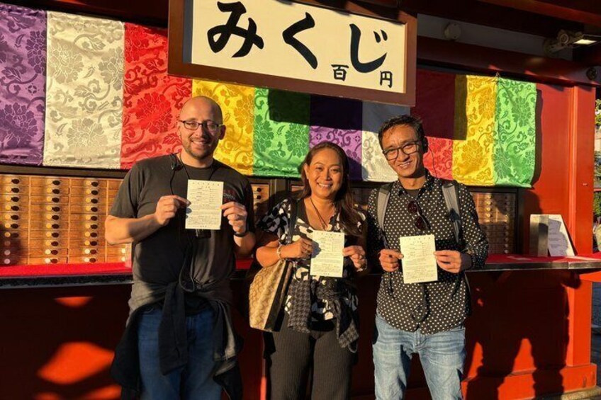 Three people holding papers in front of a colorful wall with a sign that reads 'みくじ'. 3