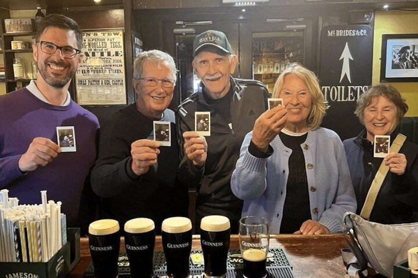 A group of people holding photographs, with Guinness beer glasses and a sign for 'McBride's & Co. Toilets' in the background. 1