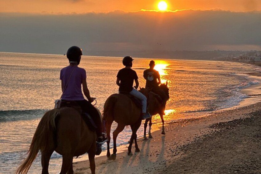 Three people riding horses along a beach at sunset. 3