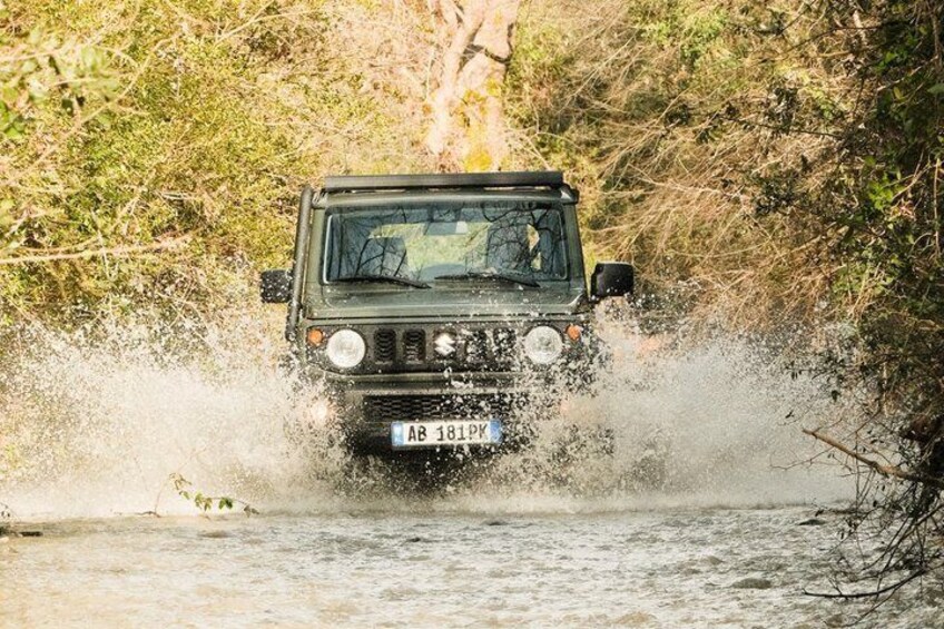 A vehicle driving through a shallow water crossing, surrounded by dense vegetation. 1