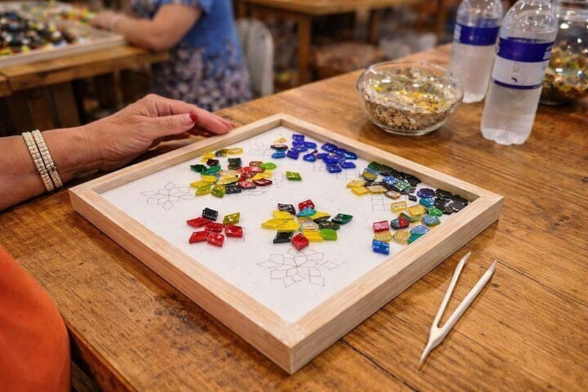 A wooden table with colorful glass pieces arranged in a pattern, a pair of tweezers, and a framed artwork. 2