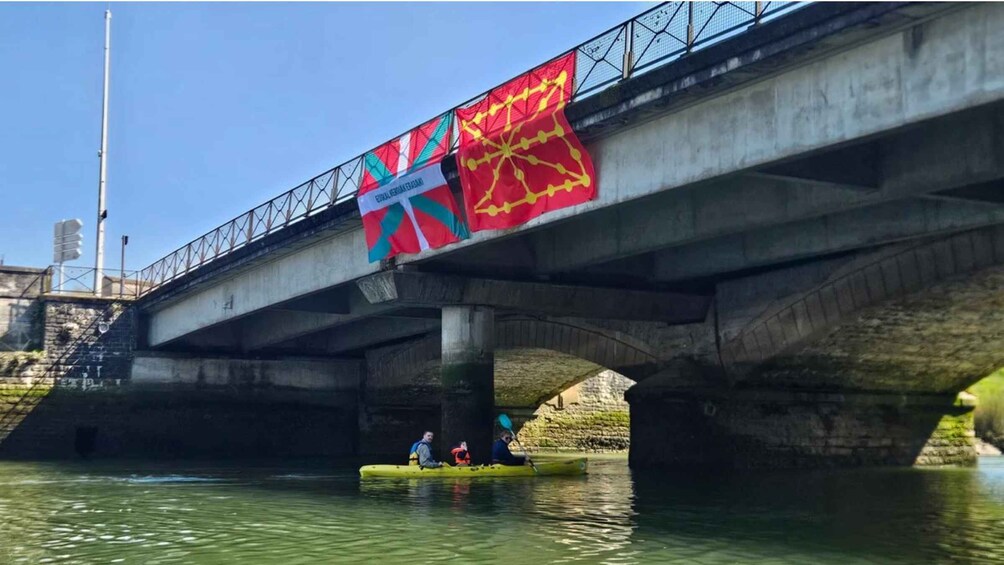 A bridge with a colorful flag, a kayak with people, and a calm river. 1