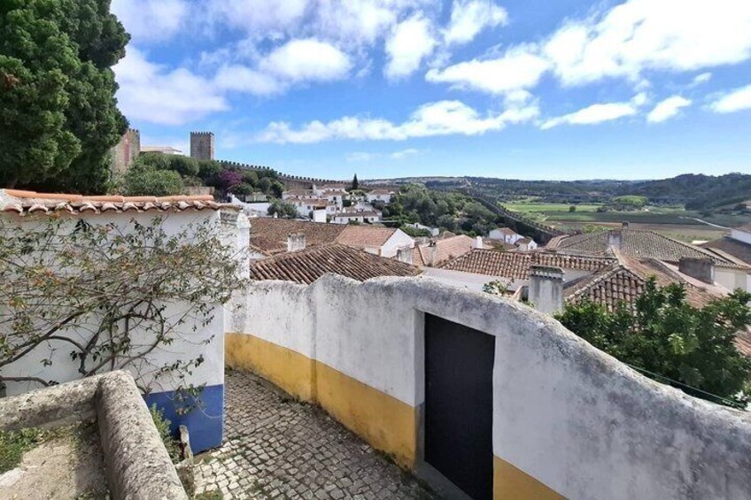 A cobblestone street with a white wall and a yellow stripe, overlooking a town with terracotta rooftops and a distant castle. 2