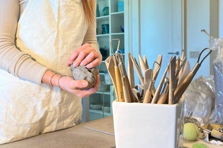 A person holding a clay object, with a container of tools and a green bowl on a table. 2
