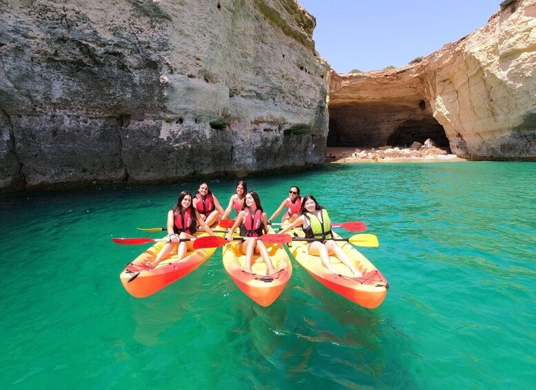 Four people in kayaks on clear turquoise water with rocky cliffs and a cave entrance in the background. 3