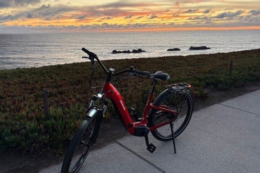 A red electric bicycle parked on a sidewalk by the sea with a sunset in the background. 2