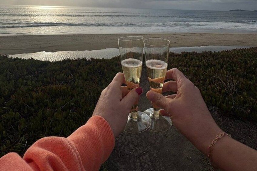 Two people toasting with glasses of beer on a beach overlooking the ocean. 3