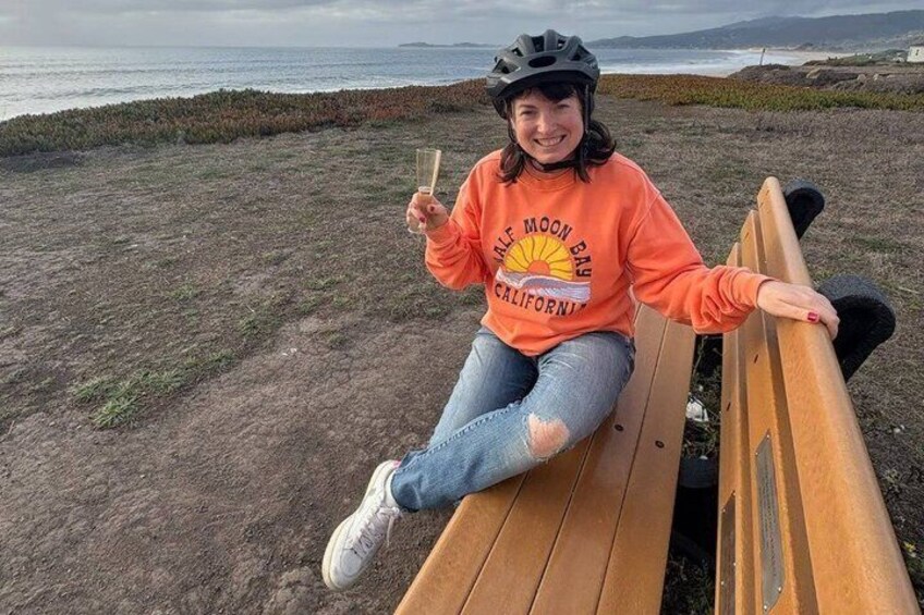 A person sitting on a wooden bench at a beach, holding a drink, with a sunset and ocean in the background. 4