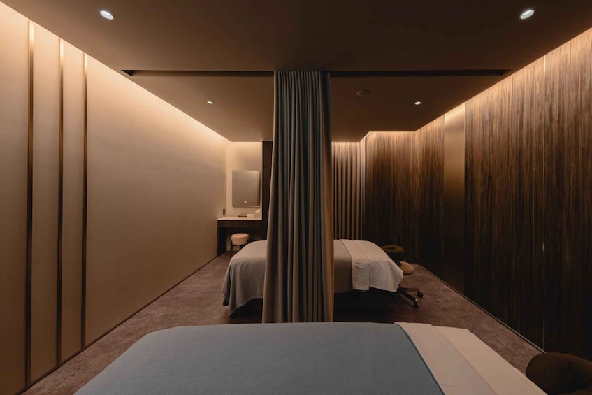 A modern spa room with a bed, two massage tables, a mirror, and wooden paneling. 2
