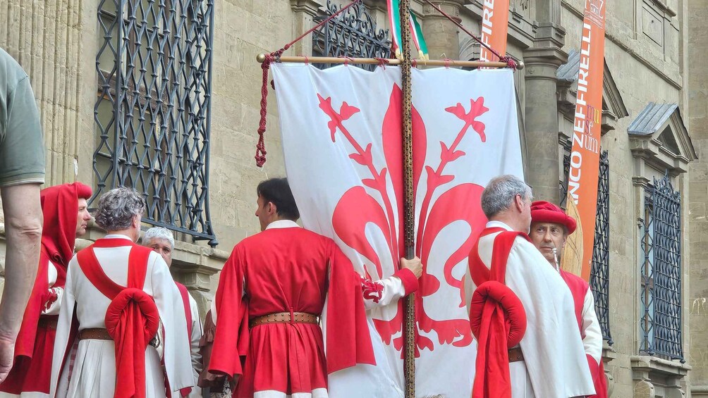 A group of individuals in red and white ceremonial attire, carrying a white banner with a red fleur-de-lis emblem, in front of a historic building with ornate ironwork. 4